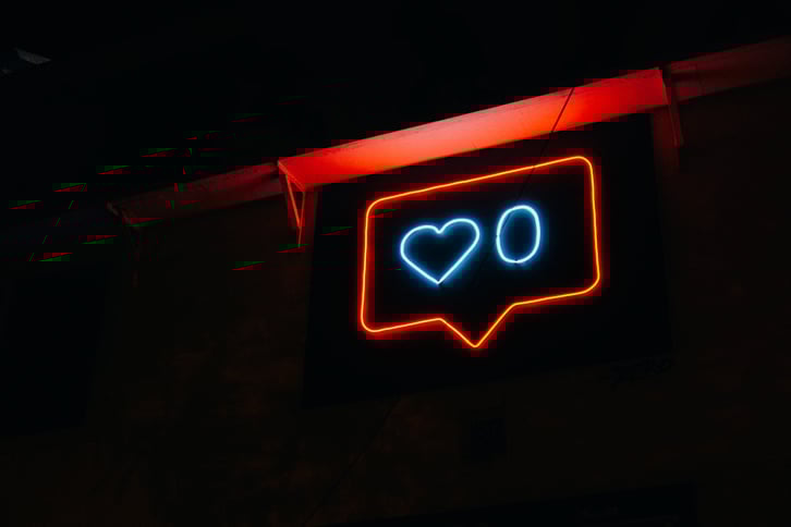 A red-and-blue lit neon sign in the darkness: a blue heart next to a zero, outlined by a red speech box. The neon sign is hanging from a red rafter.